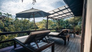 Una terraza de madera con dos sillones y una sombrilla grande, rodeada de abundante vegetación.