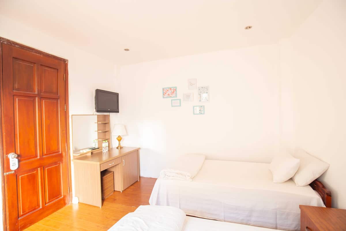 A hotel room with a wooden door, a desk with a lamp, a television, two beds, and a wall with framed pictures.