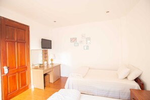 Habitación de hotel con puerta de madera, un escritorio con lámpara, televisor, dos camas y un muro con cuadros enmarcados.