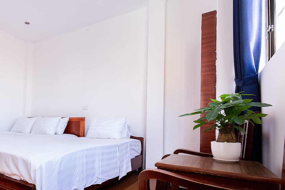 A bedroom with a bed, a wooden nightstand, a potted plant, and a window with curtains.