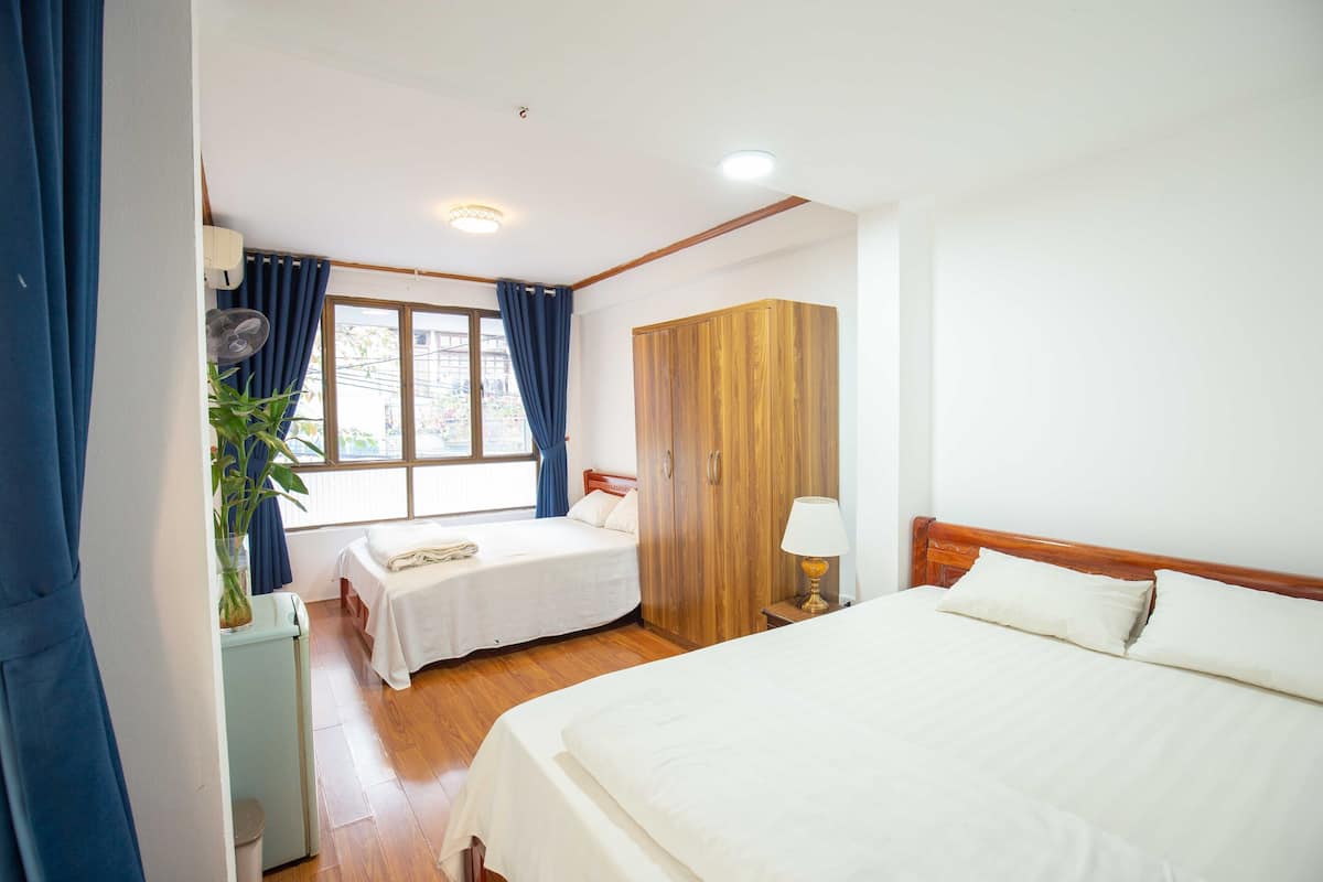 A hotel room with two beds, a wooden wardrobe, a bedside table with a lamp, a window with blue curtains, and a small plant on a nightstand.