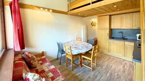 Ein gemütliches Zimmer mit Holzbalkendecke, einem Esstisch mit Stühlen, einer Couch und einer Küchenzeile mit Holzausbau und Spüle.