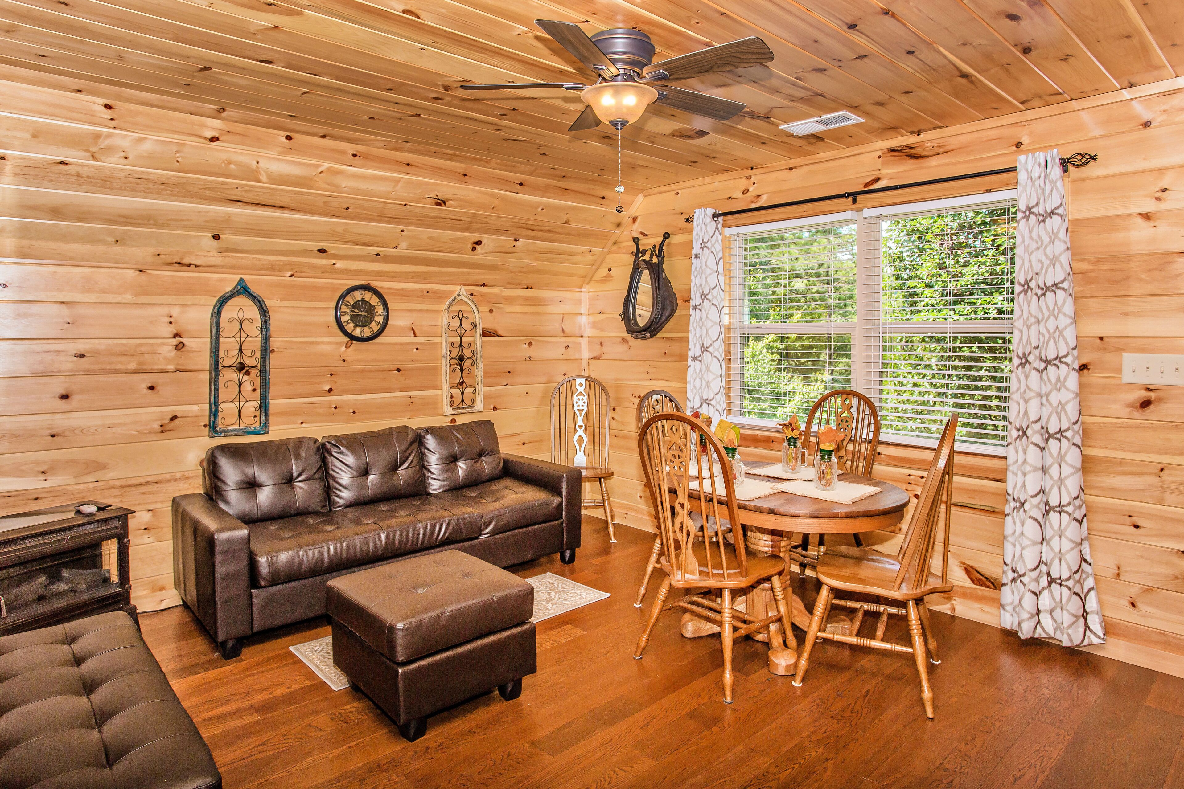 A cozy cabin interior with a wooden ceiling, a leather sofa, a dining table with chairs, and a large window with curtains.