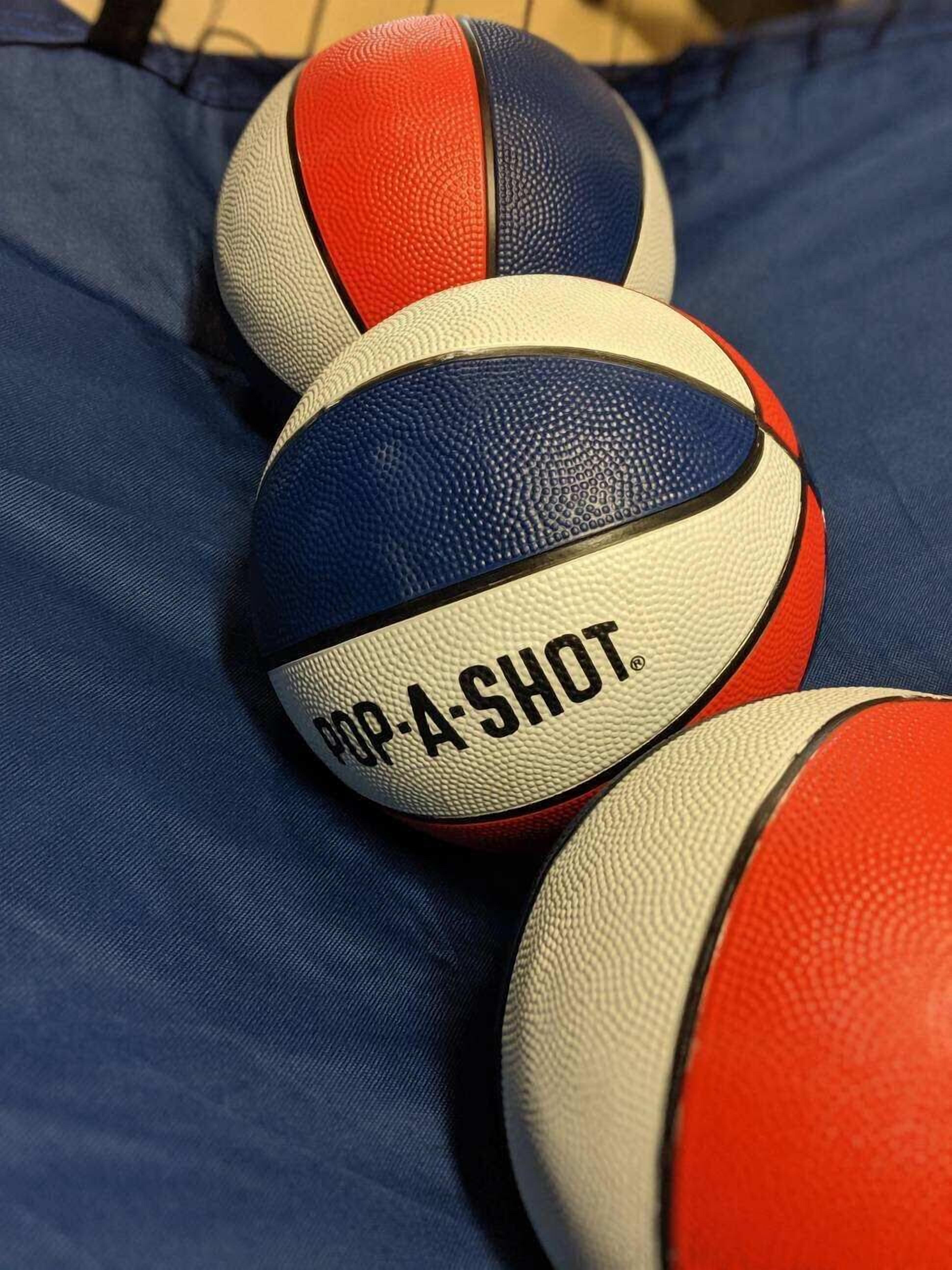 A close-up of three basketballs with the text "POP-A-SHOT" on them, placed on a blue fabric surface.