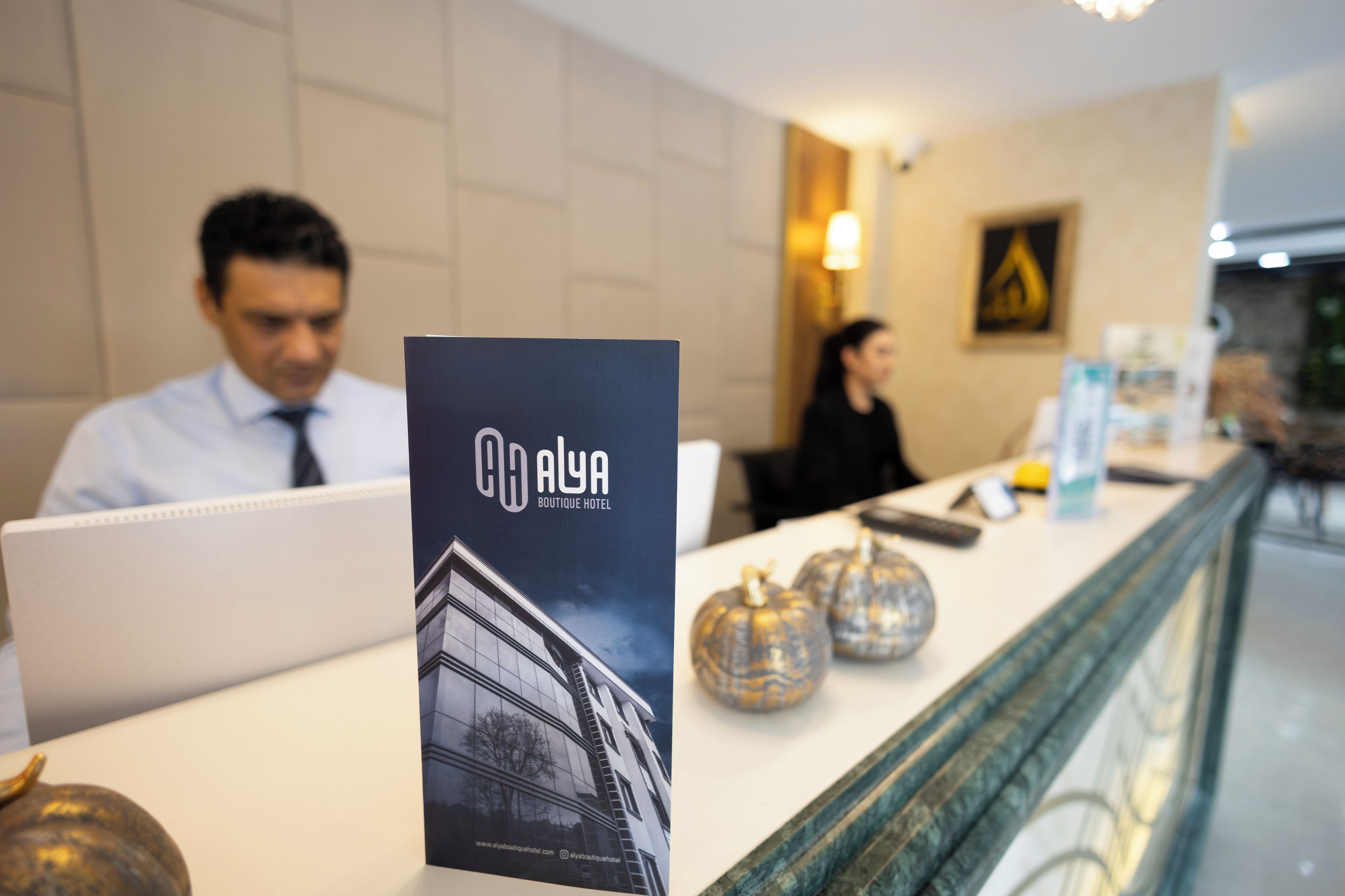 En receptionsdisk med en brochure for Alya Boutique Hotel, en bærbar computer og dekorative græskar.