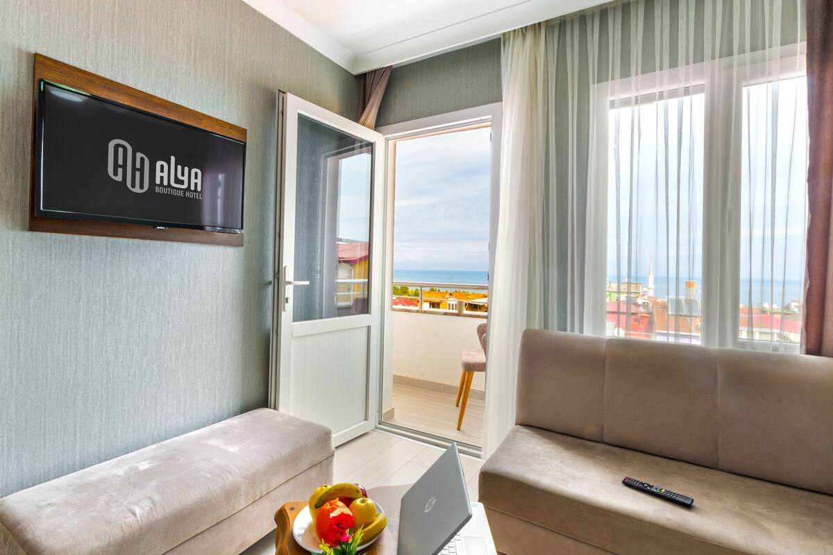A hotel room with a balcony, a sofa, a laptop, and a TV displaying the hotel name.