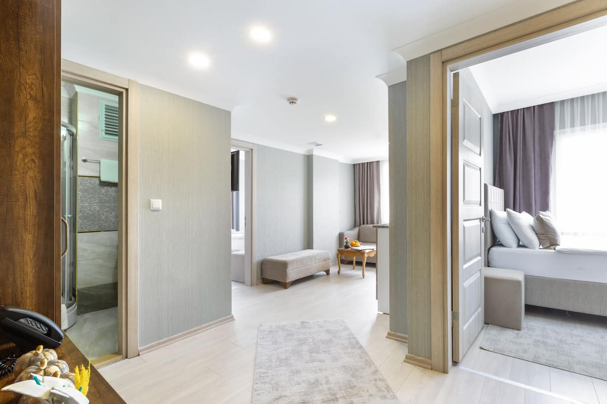 A modern hotel room with a large bed, a seating area with a sofa and armchairs, a small table with a fruit bowl, and a bathroom with a shower and toilet.