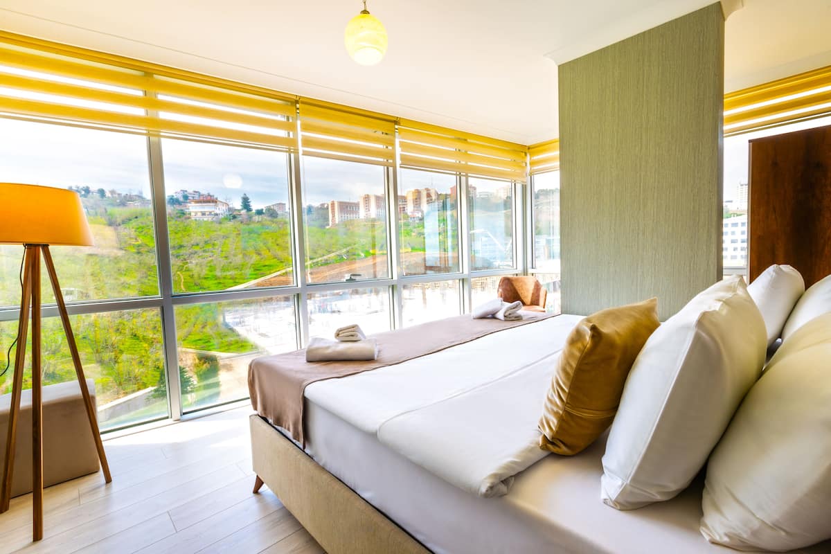 A modern hotel room with a large window, a bed with white linens, and a standing lamp.