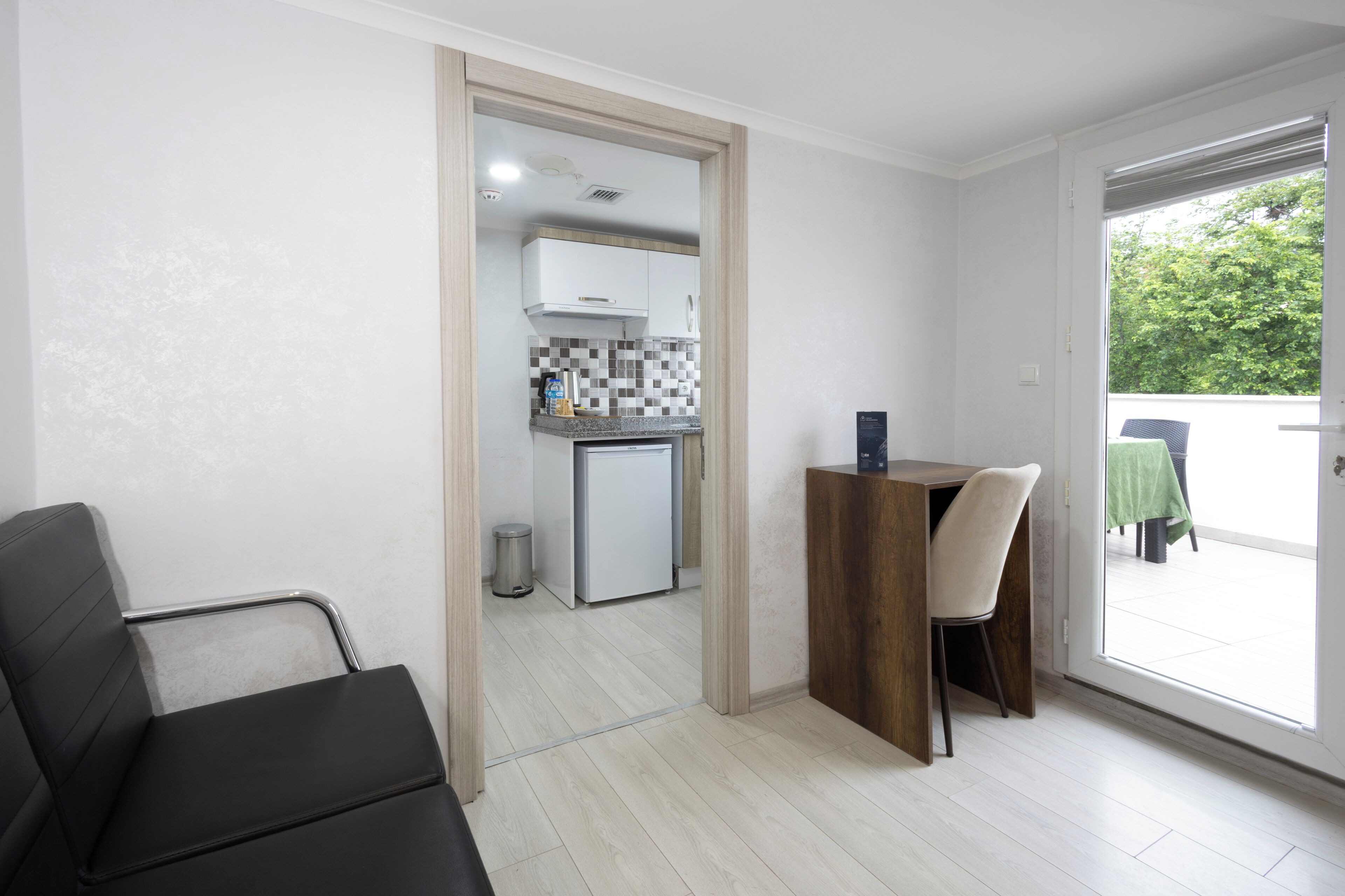 A modern kitchen with white cabinets, a tiled backsplash, and a small refrigerator. There is a dining area with a wooden table and chairs, and a large window offering a view of greenery outside.