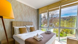 Een hotelkamer met een groot bed, een bedlampje en uitzicht op een groen landschap door grote ramen.