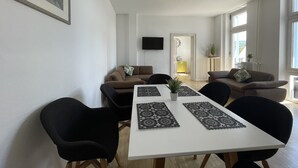 A modern living room with a dining area, featuring a white table, black chairs, and two sofas with cushions. A flat-screen TV is mounted on the wall, and a window provides natural light. - Apartment in the heart of Bad Pyrmont (Bad Pyrmont)