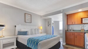 Habitación de hotel con cama, mesita de noche, lámpara y vista a la cocina.