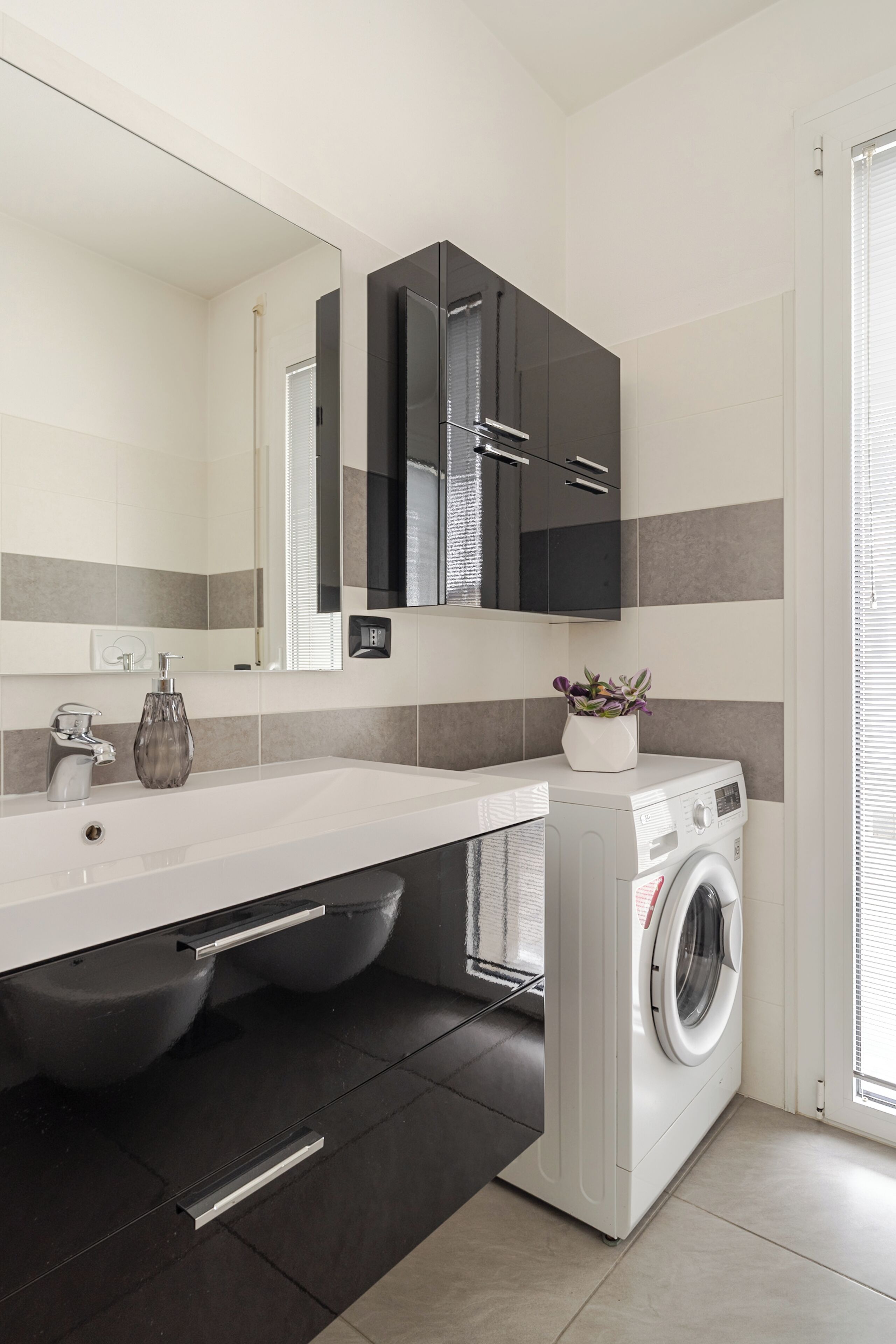 Un baño moderno con un lavamanos blanco, gabinetes negros, lavadora integrada y un espejo grande.