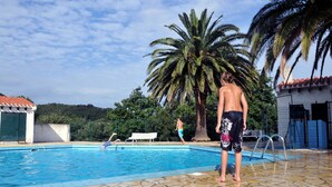 Une piscine avec une terrasse carrelée, des palmiers et un bâtiment surmonté d’un toit recouvert de tuiles rouges.