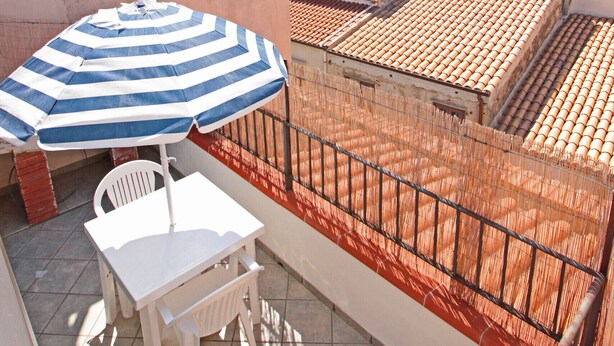 A balcony with a striped umbrella, white table, and chairs, overlooking a mountainous landscape and terracotta-roofed buildings. - Terrazza Cathedral by Wonderful Italy (Cefalù)