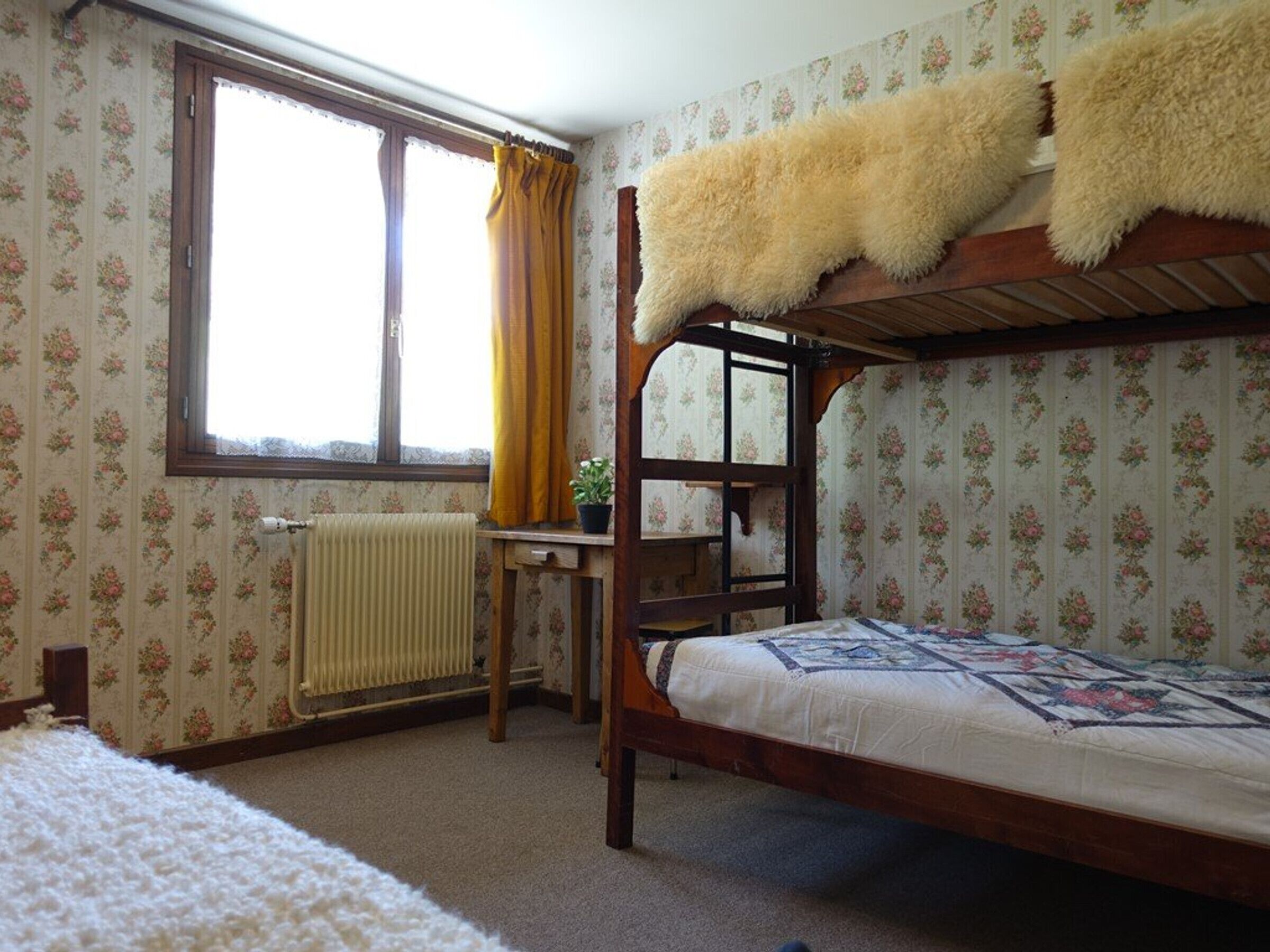 A bunk bed room with floral wallpaper, a radiator, a window with curtains, and a wooden desk.