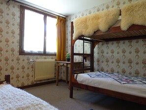 Quarto com beliche, papel de parede floral, radiador, janela com cortinas e escrivaninha de madeira.