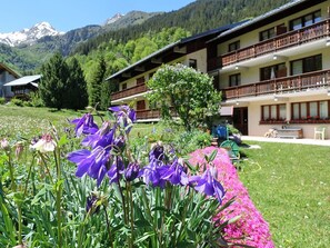 Un hotel con vistas a la montaña, un jardín de flores moradas y un balcón con barandilla de madera.