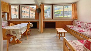 Una habitación con una mesa y sillas de madera, un sofá y un televisor montado en la pared. Amplias ventanas ofrecen vistas a edificios cercanos.