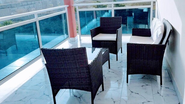 A balcony with wicker chairs and a glass table, large windows, and a tiled floor.