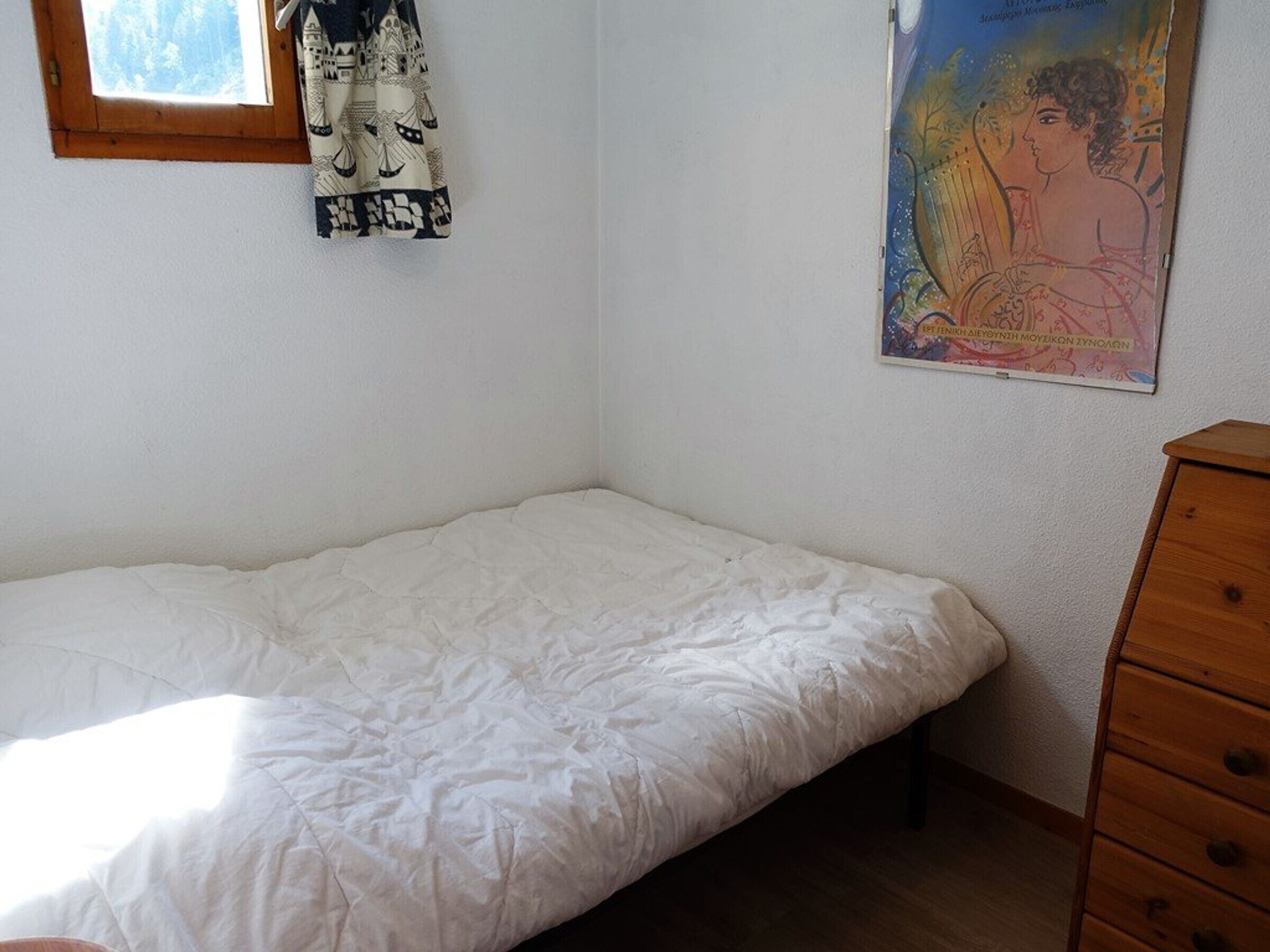 Une petite pièce, peu meublée, avec un lit simple, une commode en bois et une fenêtre.