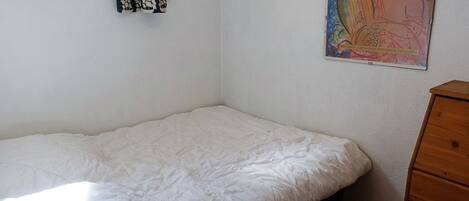 Une petite pièce, peu meublée, avec un lit simple, une commode en bois et une fenêtre.