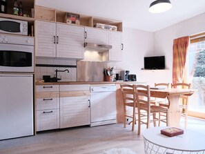 Et lille køkken med hvide skabe, et spisebord med stole, et fjernsyn og et vindue med gardiner.