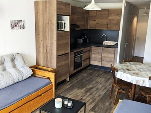 Eine kompakte Küche mit Holzausstattung, einer Mikrowelle und einer Kaffeemaschine. Ein Esstisch mit weißem Tischlaufzeug und Holzstühle. Ein graues Sofa lehnt an der Wand, und ein kleiner Tisch mit Kerze und Gläsern steht im Vordergrund.