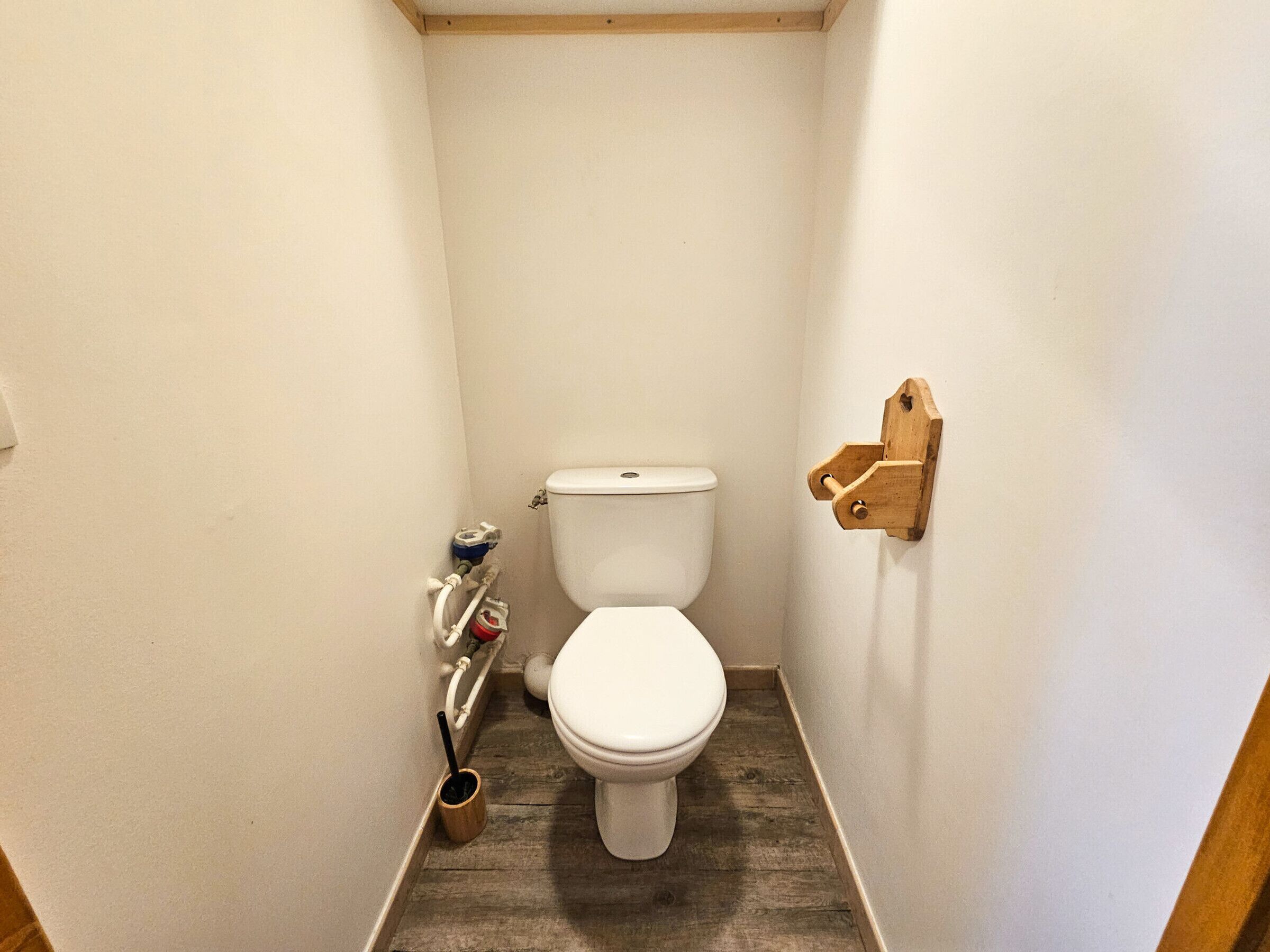 A small, narrow bathroom with a white toilet, wooden floor, and a wooden shelf on the wall.