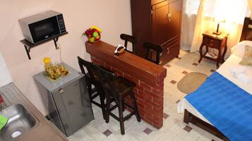 Een compacte hotelkamer met een kitchenette, een bed met een blauwe deken, een kleine tafel met stoelen, een magnetron, een koelkast en een schaal fruit.