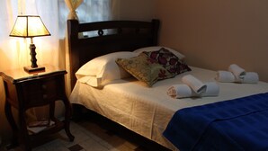 Een net opgemaakt bed met kussens en een opgevouwen handdoek, een bedlampje en een klein houten nachtkastje met een boek en een afstandsbediening.