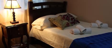Cama bem arrumada com almofadas e uma toalha dobrada, abajur e mesa de cabeceira de madeira pequena com um livro e um controle remoto.