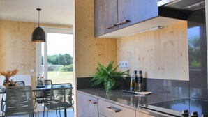 A modern kitchen with wooden cabinets, a black countertop, and a stainless steel stove. There is a dining area with a table and chairs, and a hanging light fixture. - Holiday home Callants` Lodge (Callantsoog)