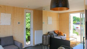 A modern room with wooden walls, a grey sofa, a black TV stand, and a large window with a view of greenery and parked cars. - Holiday home Callants` Lodge (Callantsoog)