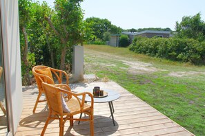 Une terrasse en bois, agrémentée de chaises en osier et d’une petite table, d’où l’on peut admirer une pelouse et un bâtiment situé au loin.