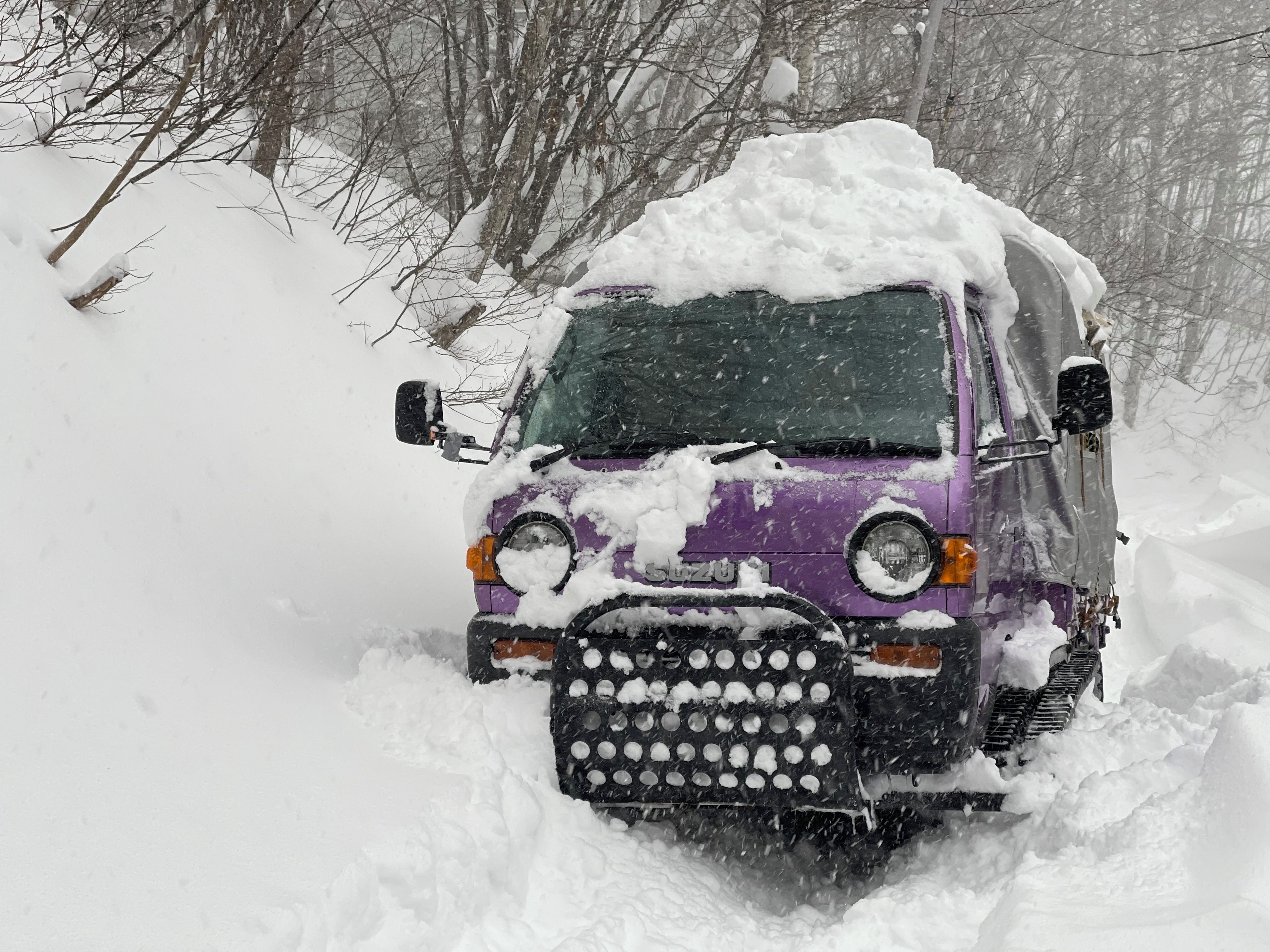 A vehicle covered in snow, parked in a snowy area with trees in the background.