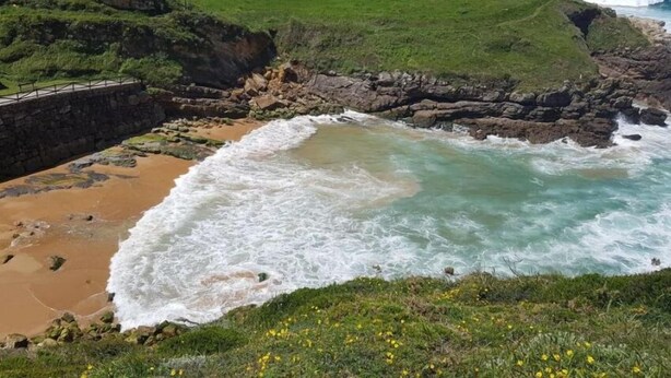 A coastal landscape with a grassy hill, rocky shoreline, and waves crashing onto the beach.