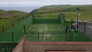 A tennis court with a green fence, surrounded by a brick wall and a grassy landscape with hills and a view of the sea.