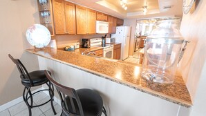 A kitchen with wooden cabinets, a granite countertop, a microwave, a stove, and two bar stools. - Oceanfront Corner Unit 2BR Condo 5th Floor Balcony Views, 501 (Indian Harbour Beach)
