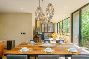 A dining area with a wooden table set for four, a modern chandelier, and large windows offering a view of greenery. - Arthouse w/ Private Pool, Gym, and Wellness Spa (Tulum)
