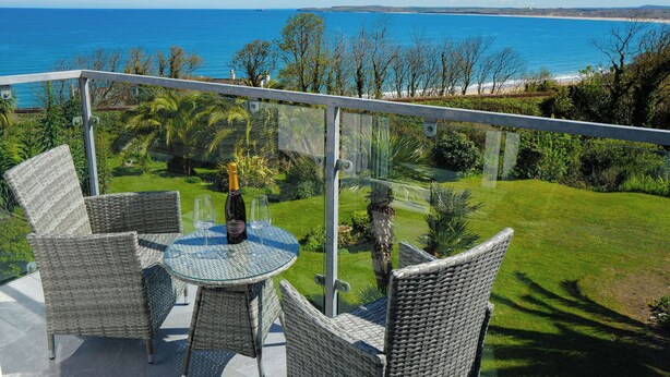Un balcón con muebles de mimbre y una mesa de vidrio, con vistas a un paisaje costero con playa y mar azul cristalino.