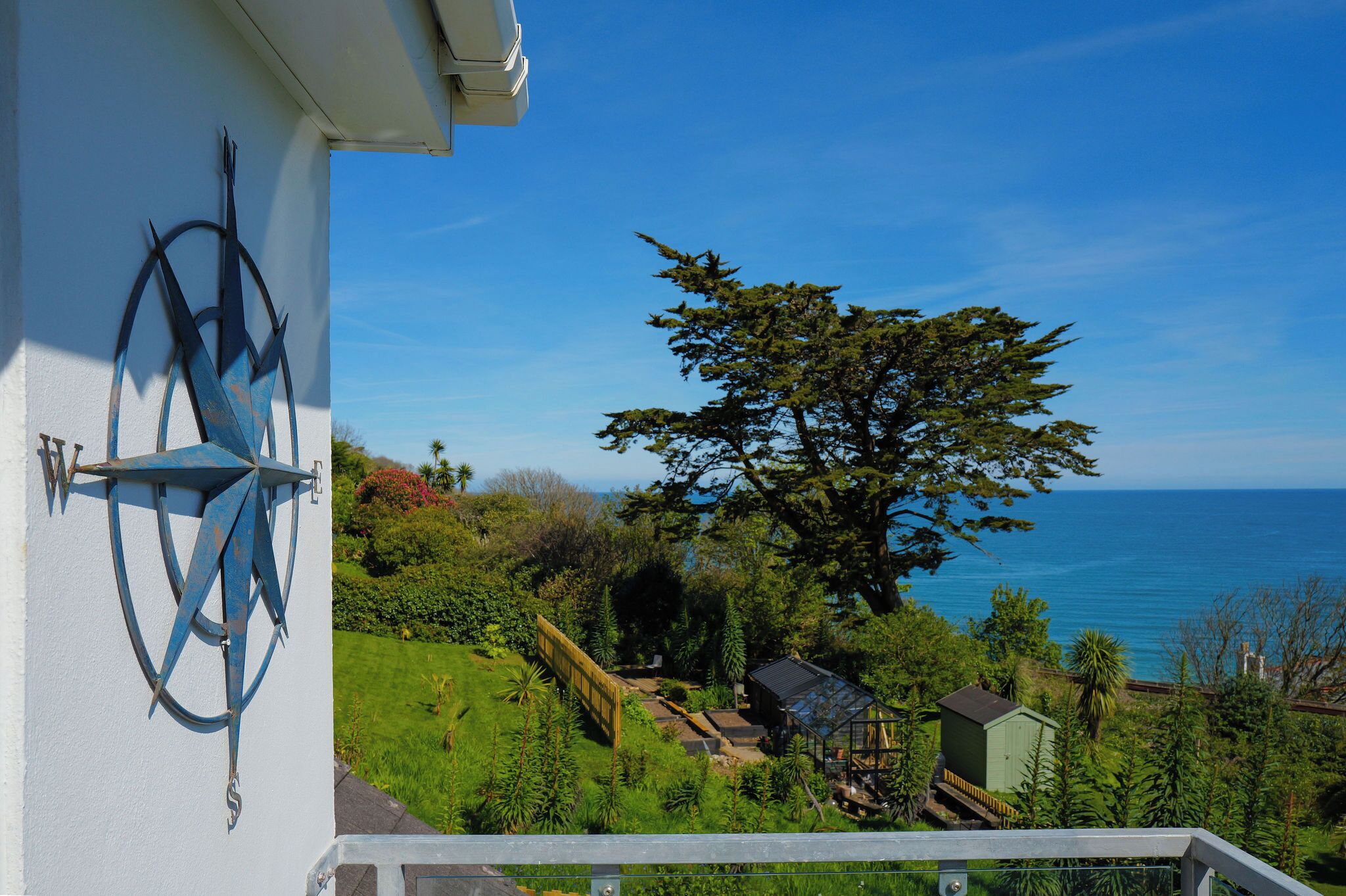 Un balcon offrant une vue sur un paysage côtier, avec un grand arbre et un ciel bleu dégagé.