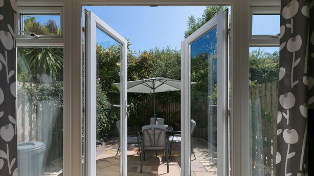 An open glass door leading to an outdoor patio with a table and chairs, a patio umbrella, and a view of a garden. - Honeysuckle Cottage, Lelant – Sleeps - On-Site Parking – Dog Friendly (Lelant)