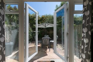 Une porte vitrée ouverte donne sur une terrasse extérieure équipée d’une table et de chaises, d’un parasol et offrant une vue sur un jardin.
