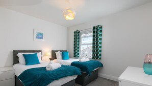 A hotel room with two beds, a window with patterned curtains, a bedside table, and a wall-mounted light fixture. - Honeysuckle Cottage, Lelant – Sleeps - On-Site Parking – Dog Friendly (Lelant)