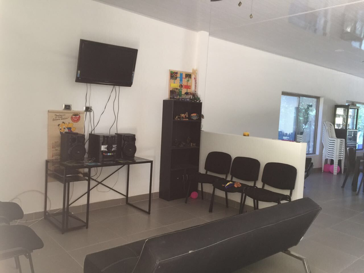 Una habitación moderna con televisor de pantalla plana montado en la pared, un mueble negro para entretenimiento con dispositivos electrónicos, una pequeña mesa con un altavoz y una zona de estar con sillas.