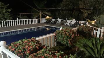 A nighttime pool area with a white balustrade, lounge chairs, and potted plants.
