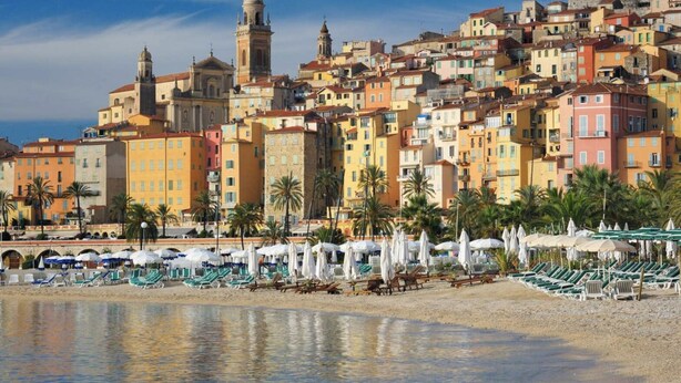 A coastal cityscape with colorful buildings, a prominent church with a bell tower, and a beach area with sun loungers and umbrellas. - Panoramic views of Monaco and the Mediterranean (Roquebrune-Cap-Martin)