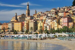 A coastal cityscape with colorful buildings, a prominent church with a bell tower, and a beach area with sun loungers and umbrellas.