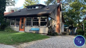 A two-story cottage with a red door, large windows, and a blue bench outside. - Big DECK Cottage, 3-min walk to BEACH- 4 bdrms, 1.5 baths 7 beds, parking 3 cars (Grand Bend)